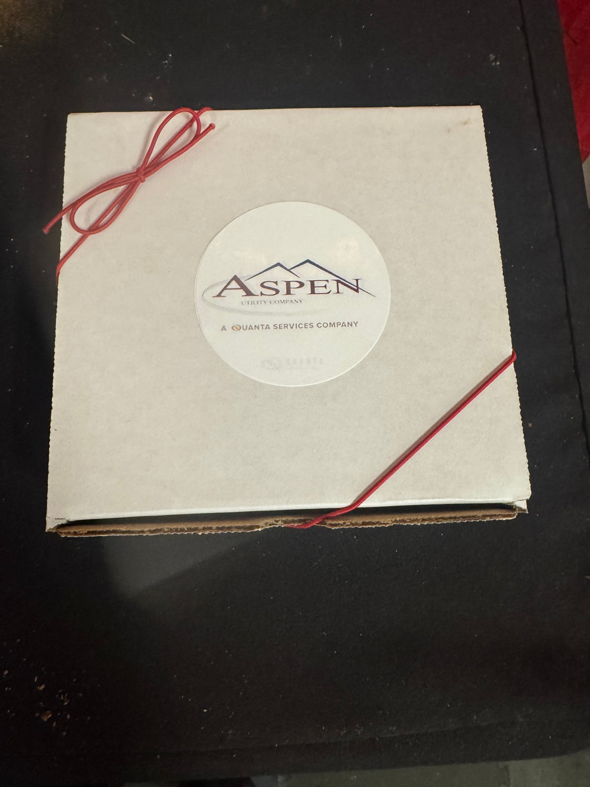 A square white cardboard box with a circular white sticker in the center reading 'ASPEN' and a red string tied through the corners, placed on a dark surface.